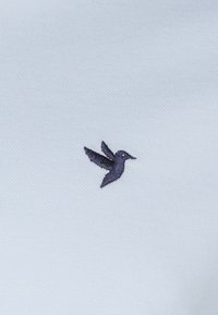 Lys blå tekstil med et mørklilla broderet kolibri-logo, der har detaljerede vinger og et distinkt buet næb, på en glat overflade.
