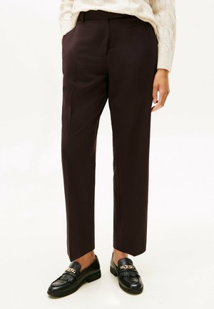 Person iført mørkebrune ankellange bukser, hvid sweater og sorte læder loafers med gyldent kædedetalje.
