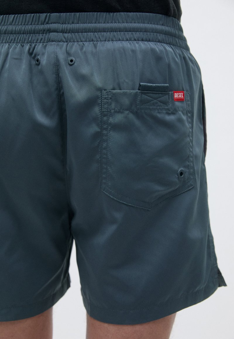 Marineblauwe, lichte zwembroek van glanzende stof. Heeft een elastische tailleband, een achterzak en een klein rood Diesel-logo.