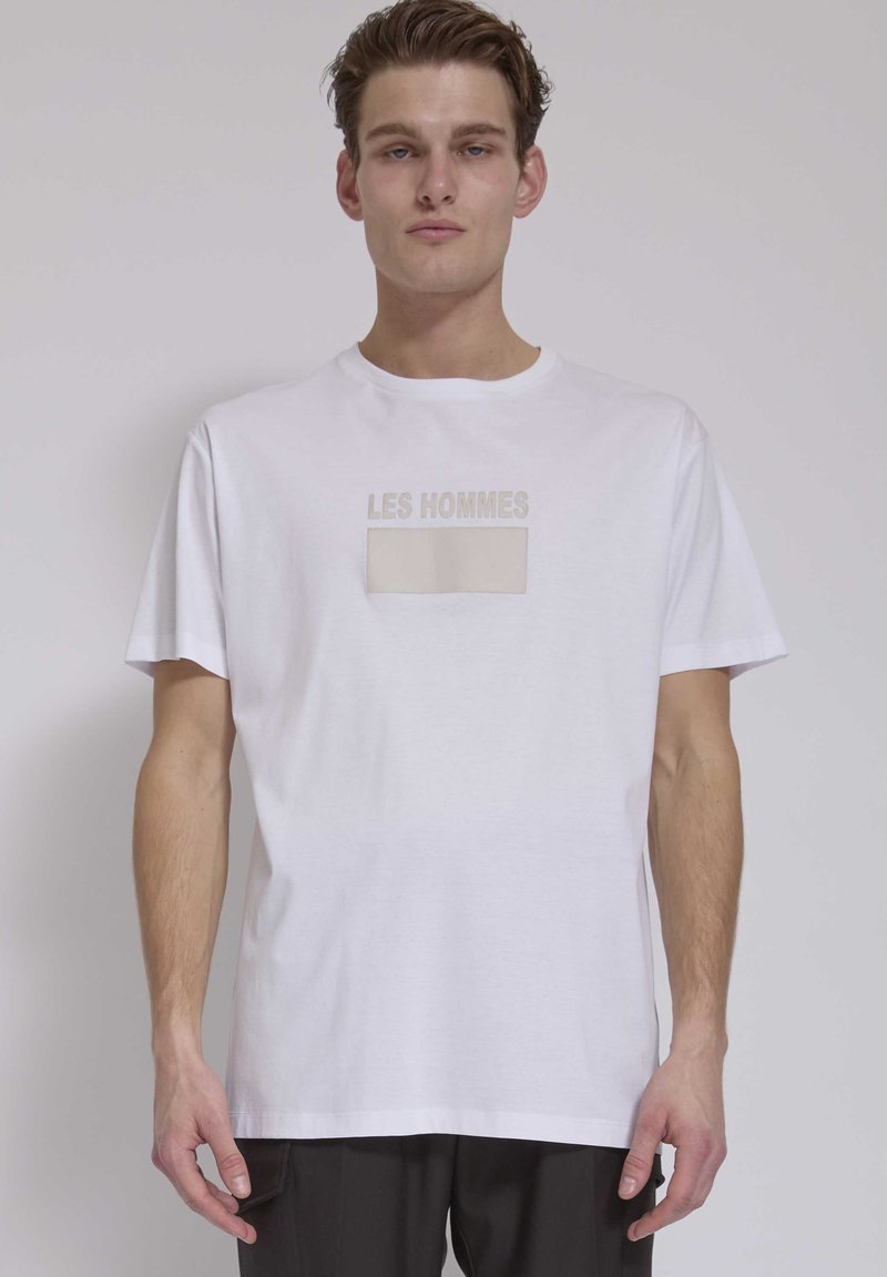 Jonge man die een wit T-shirt draagt met beige tekst "LES HOMMES" en een leeg beige rechthoekje op de borst, staand tegen een effen achtergrond.