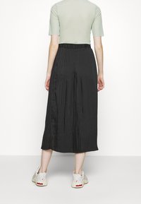 Černá plisé midi sukně s elastickým pasem, kombinovaná s přiléhavým zeleným topem a bílými teniskami, zobrazená zezadu.