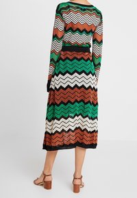 Kvinna i en långärmad klänning med zickzackmönster i grön, orange, svart och vit, tillsammans med bruna klacksandaler, stående med ryggen mot kameran.