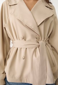 Veste beige croisée avec grand col, ceinture en tissu et boutons, présentant une texture douce et des détails plissés à la taille.