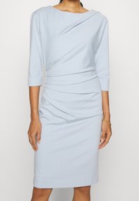 Vestido azul claro de tejido suave, con mangas tres cuartos, un diseño drapeado y una silueta ajustada que se ciñe en la cintura.