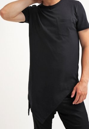 Homme portant un t-shirt noir à manches courtes avec un ourlet asymétrique et une poche poitrine, posant avec un bras levé derrière la tête.