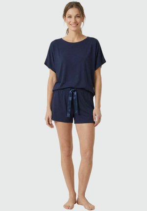 Vrouw glimlacht, staat op blote voeten en draagt een marineblauw kort mouwtopje en bijpassende shorts met een satijnen trekkoord op een effen achtergrond.