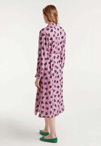 Vestido rosa con estampado de fresas en negro y verde, mangas largas, cintura ceñida con cinturón y largo a la mitad de la pantorrilla. Zapatos planos verdes complementan el conjunto.
