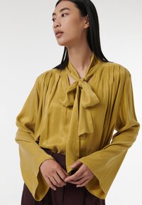Blusa mostaza con cuello atado, tela texturada, hombros plisados y mangas acampanadas, combinada con una falda oscura.