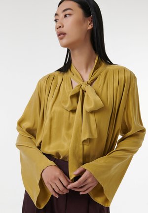 Mosterdgele blouse met een strik rond de hals, gemêleerde stof, geplooide schouders en klokmouwen, gedragen met een donkere rok.