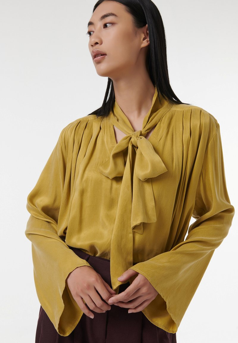 Blusa mostaza con cuello atado, tela texturada, hombros plisados y mangas acampanadas, combinada con una falda oscura.