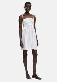Weißes ärmelloses Kleid mit gerafftem Bund und Kordelzugdetail, aus weichem Stoff, reicht bis über die Knie, kombiniert mit schwarzen Schuhen.