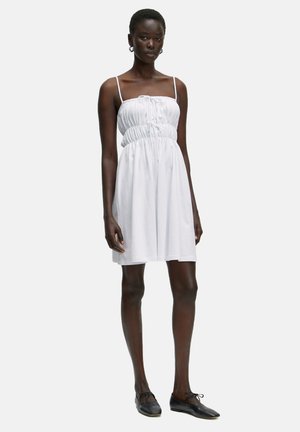 Weißes ärmelloses Kleid mit gerafftem Bund und Kordelzugdetail, aus weichem Stoff, reicht bis über die Knie, kombiniert mit schwarzen Schuhen.