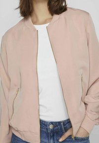 Light pink bomber jacket with a smooth texture, gold zipper, ribbed collar, and cuffs. Worn over a white shirt and paired with blue jeans.