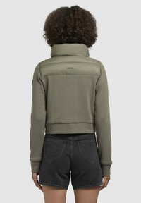 Sweatshirt scurt din material verde oliv cu guler pufos, asortat cu pantaloni scurți din denim închis. Prezintă logo pe guler și un design texturat.