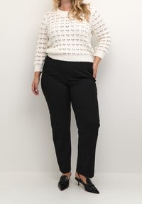 Weißer Strickpullover mit offenem Webmuster, kombiniert mit schwarzen maßgeschneiderten Hosen. Schwarze spitz zulaufende High Heels runden das Outfit ab.
