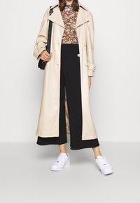 Beige trenchcoat med stora slag, blommönstrad topp, svarta vida byxor och vita sneakers med texturerade detaljer. Svart axelremsväska.