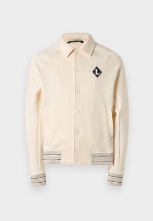 Εκρού σακάκι με κουμπιά και γιακά, δύο πλαϊνές τσέπες, πλεκτά μανίκια και τελείωμα με ρίγες, καθώς και μαύρο διαμαντένιο λογότυπο στο στήθος.