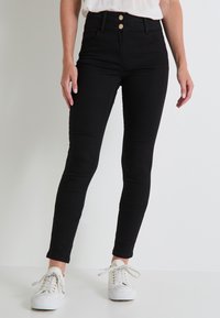 Jeans negros de cintura alta con dos botones en tono dorado, diseño ajustado y a la altura del tobillo, combinados con zapatillas blancas. Textura suave.