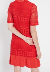 Červené krajkové šaty s krátkými rukávy, které mají strukturovaný vzor. Sukně má volánkový lem, což kontrastuje s krajkovým tělem.