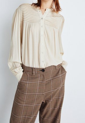 Mujer con una blusa de botones plisada color crema metida en un pantalón de cintura alta de cuadros marrones, con las manos en los bolsillos, frente a un fondo blanco.