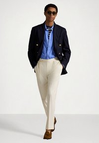 Μπλε διπλόμπρεσο σακάκι του ναυτικού, ανοιχτό γαλάζιο πουκάμισο με κουμπιά, λευκό παντελόνι και καφέ μοκασίνια. Το μοντέλο στέκεται με τα χέρια χαλαρά στα πλάγια.