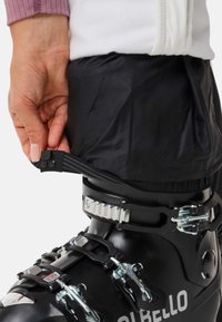 Zwarte skischoen met metalen gespen en riem, gecombineerd met een strakke zwarte broekspijp die vastzit met een drukknoopsluiting. Hand die de broekspijp vasthoudt.