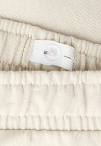 Kremowa tkanina o prążkowanej fakturze. Elastyczny pas z białą metką przymocowaną plastikowym guzikiem. Gładki, miękki materiał - szczegół.