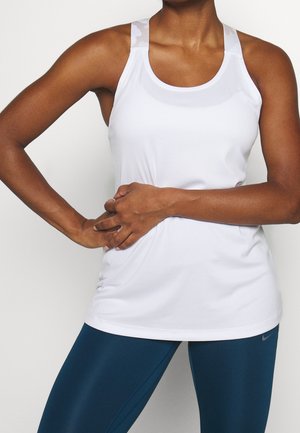 Personne athlétique portant un débardeur blanc et des leggings bleu foncé, posant avec une main sur la hanche et l'autre sur l'abdomen.