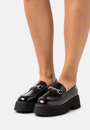 Black patent leather loafers with gold buckle detail, featuring a chunky black platform sole and textured tread for traction.