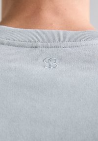 Světle šedá ribbená tkanina s detailním pohledem na límec. Na zadní straně se nachází vyšívané logo ve stejném barevném odstínu niti.