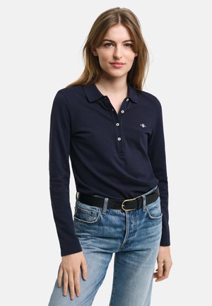 Polo a maniche lunghe blu navy con colletto con bottoni, caratterizzato da un piccolo logo e una trama liscia, abbinato a jeans in denim azzurro chiaro e una cintura nera.