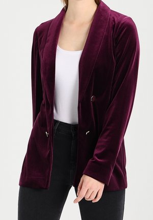 Femme portant un blazer en velours violet foncé sur un top blanc et un pantalon noir, la main détendue le long du corps.