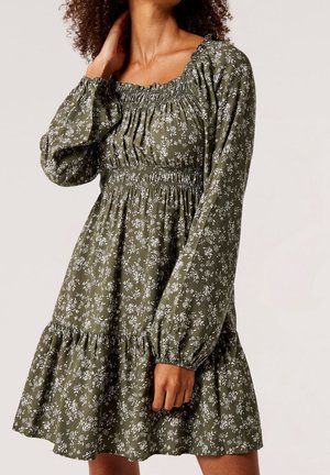 Femme portant une robe longue à manches longues olive à motifs floraux, avec un corsage smocké et une jupe plissée, posant avec une main touchant son cou.