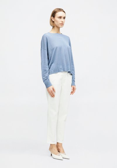 Γυναίκα που στέκεται στραμμένη ελαφρώς προς τα δεξιά, φορώντας ένα φαρδύ ανοιχτό μπλε πουλόβερ, λευκό παντελόνι και λευκά ψηλοτάκουνα παπούτσια με μυτερή μύτη, μπροστά από ένα απλό φόντο.