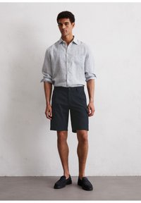 Camisa de manga larga a rayas azul claro y blanca, pantalones cortos negros y mocasines negros, de pie contra una pared neutra con superficie texturizada.