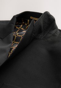 Sort blazer med et elegant design, tekstureret krave og foret med et mønster af forbundne cirkler i guld, rød og brun.