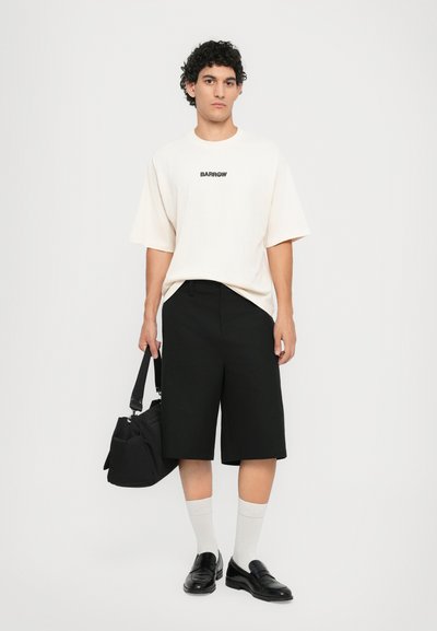 T-shirt en coton beige avec logo noir, associé à un short noir jusqu'aux genoux. Le modèle porte des chaussettes grises et des mocassins noirs, tenant un sac noir.