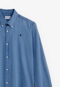 Blaues, langärmliges Hemd mit Knöpfen, knöpfbarer Kragen und kleinem, dunklem Totenkopf- und Knochenkreuz-Logo auf der linken Brust.