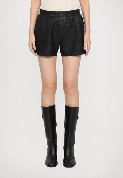Schwarze Lederkurzshorts mit einem elastischen Bund, die eine zerknitterte Textur und einen lockeren Sitz aufweisen. Kombiniert mit hohen, schwarzen, glänzenden Stiefeln.