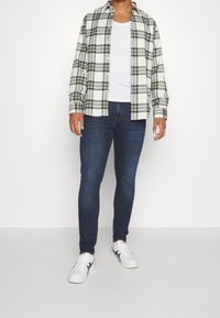 Rutigt flanellskjorta i vitt, grönt och marinblått, över en vit tanktop; parat med mörkblå jeans och vita sneakers.