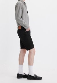 Camisa de botón gris, pantalones cortos de mezclilla negros y mocasines negros con calcetas blancas. Los detalles incluyen un parche de cuero en los pantalones cortos y dobladillos enrollados.