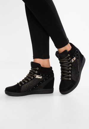 Czarne sneakersy na koturnie z złotymi oczkami i klamrami, noszone z czarnymi legginsami na białym tle.