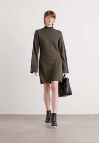 Ελιάς πράσινο πλεχτό φόρεμα με ψηλό γιακά, μακριά μανίκια, πλαϊνές σχισμές, συνδυασμένο με μια υφασμάτινη μαύρη τσάντα και μποτάκια μέχρι τον αστράγαλο.