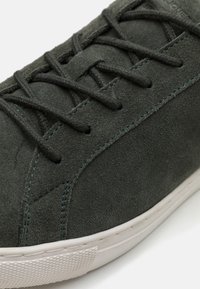 Zapatilla de ante gris con cordones planos, costuras en teal contrastante y una suela de goma blanca texturada. Presenta un diseño liso y aerodinámico.