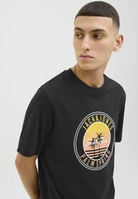 Camiseta de algodón negra con un gráfico circular que presenta palmeras, colores del atardecer y el texto "JACK & JONES PALM SPRING" en colores contrastantes.