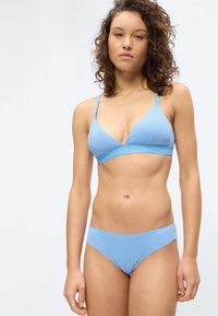 Νεαρή γυναίκα με σγουρά μαλλιά, φοράει γαλάζιο σουτιέν και συγχρονισμένο εσώρουχο, στέκεται μπροστά σε ένα απλό λευκό φόντο.