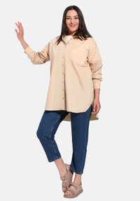 Camicia beige con bottoni e tasca sul petto, abbinata a jeans blu a maniche corte e sandali taupe con design a fascia.