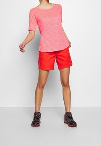 Camisa de manga corta rosa con un patrón floral texturizado, combinada con shorts naranja brillante. Un calzado negro y rojo completa el conjunto.