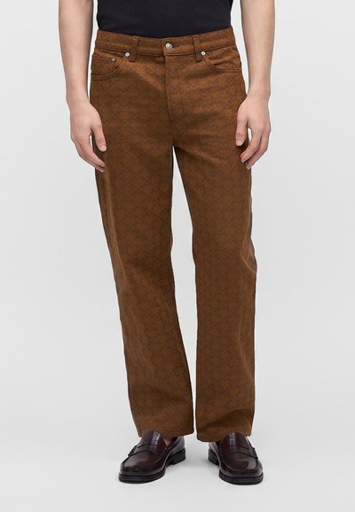 Homme portant un pantalon droit marron à motifs et des mocassins en cuir marron foncé, debout devant un fond uni.