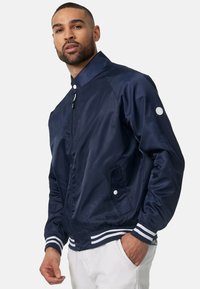 Giacca bomber blu navy con una finitura liscia e lucida, polsini a righe bianche e dettagli con bottoni a pressione. Caratterizzata da tasche laterali e un design aderente.